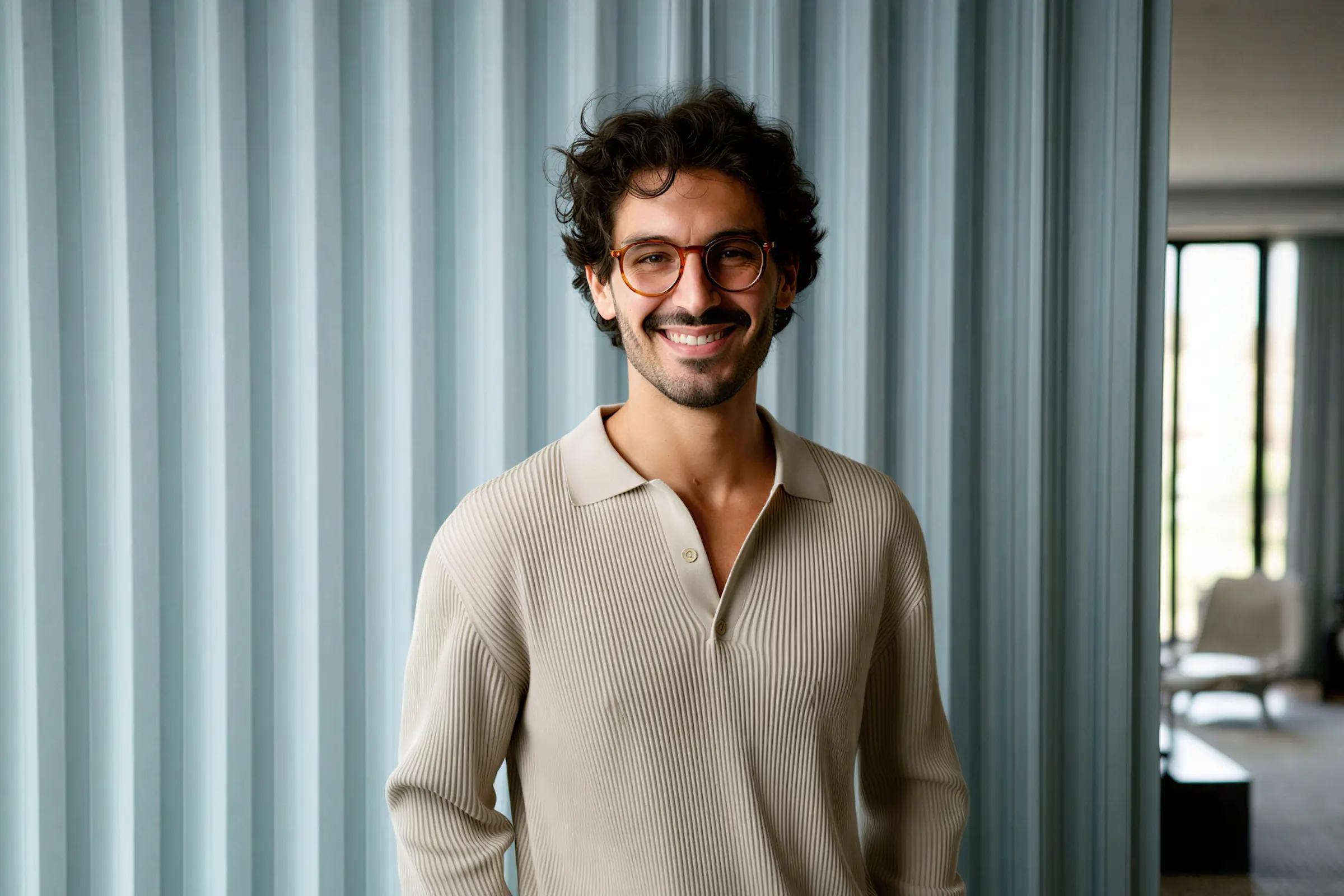 Smiling man with curly dark hair, beard, and glasses wearing a beige ribbed polo shirt in front of a blue curtain.